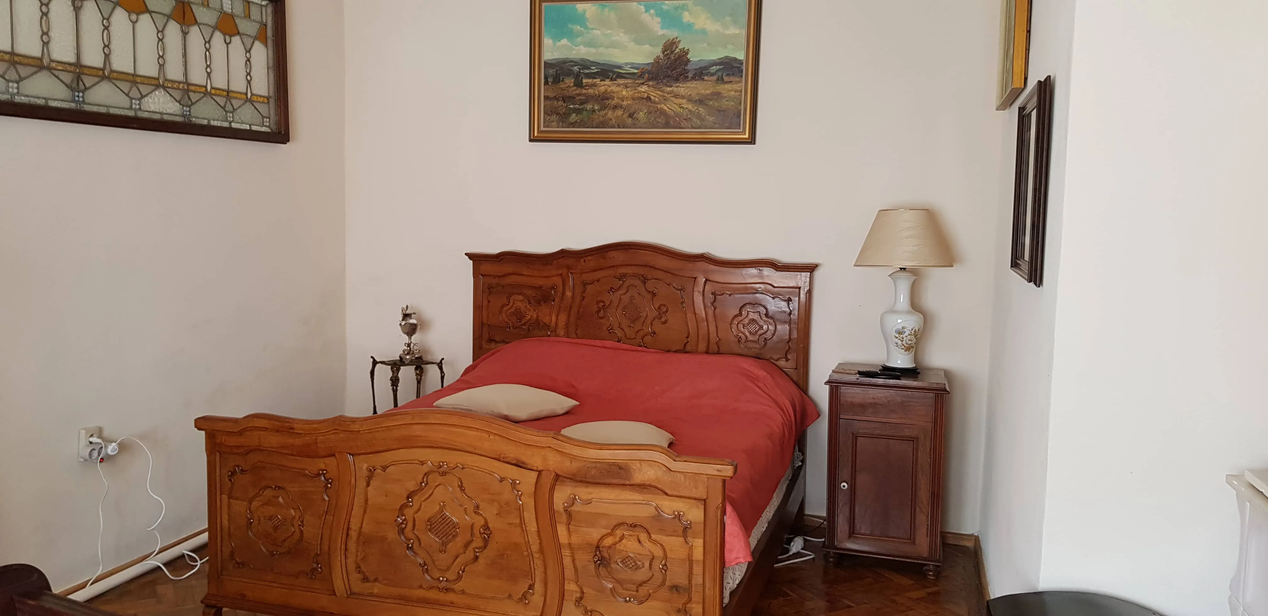 Cozy bedroom with a carved wooden bed frame covered by a red blanket and two beige pillows, a wooden nightstand with a white floral lamp, a small table with a decorative item beside the bed, and framed paintings on white walls.
