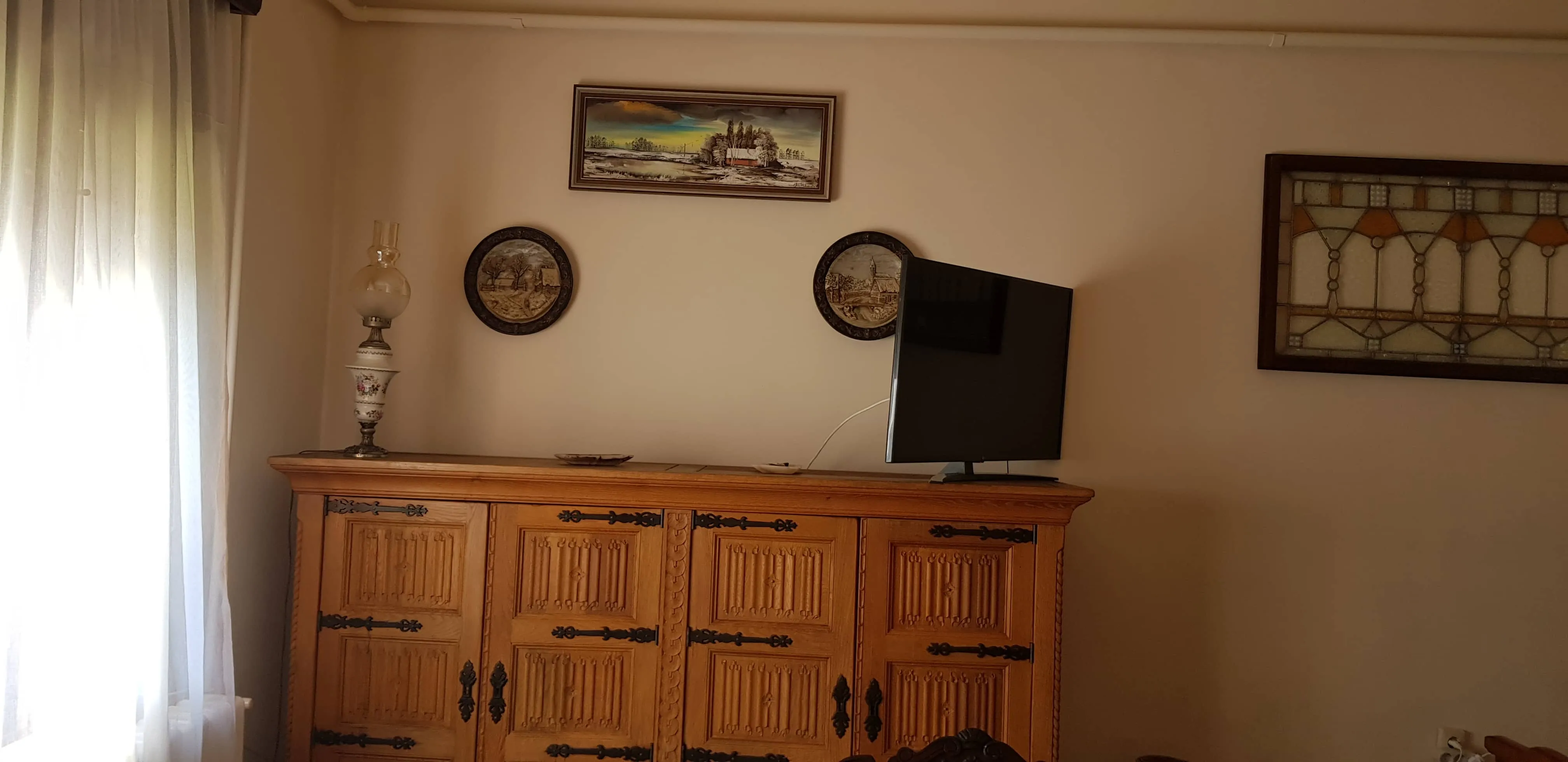 Living room corner with a wooden carved cabinet topped with a decorative lamp and a flat-screen TV, beige walls with three framed artworks, and sheer white curtains on the left window.
