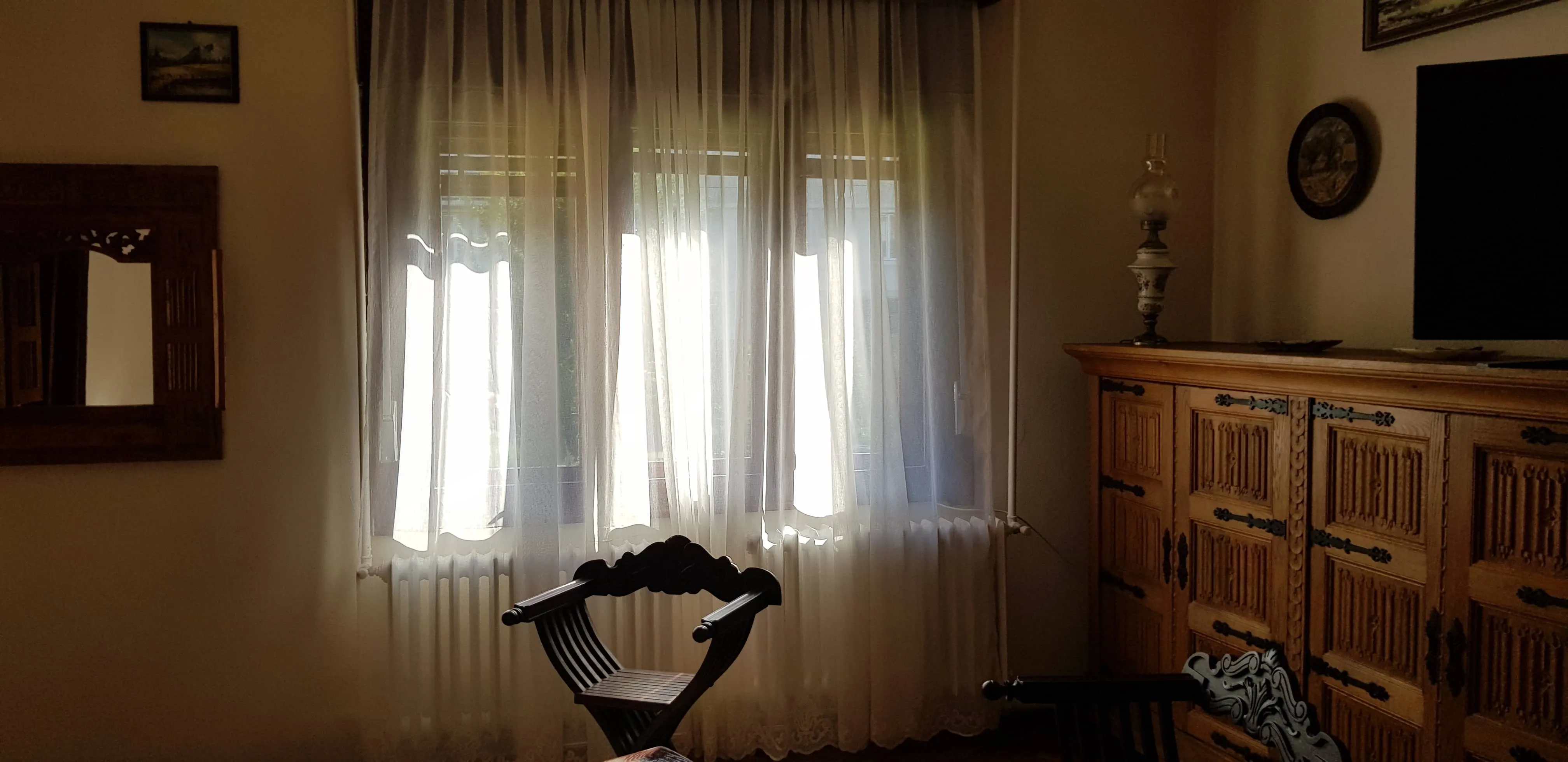 Room with sheer white curtains covering a window, a dark wooden chair with carved backrest in front of the window, and an ornate wooden cabinet on the right with a decorative lamp and a flat-screen TV above it.