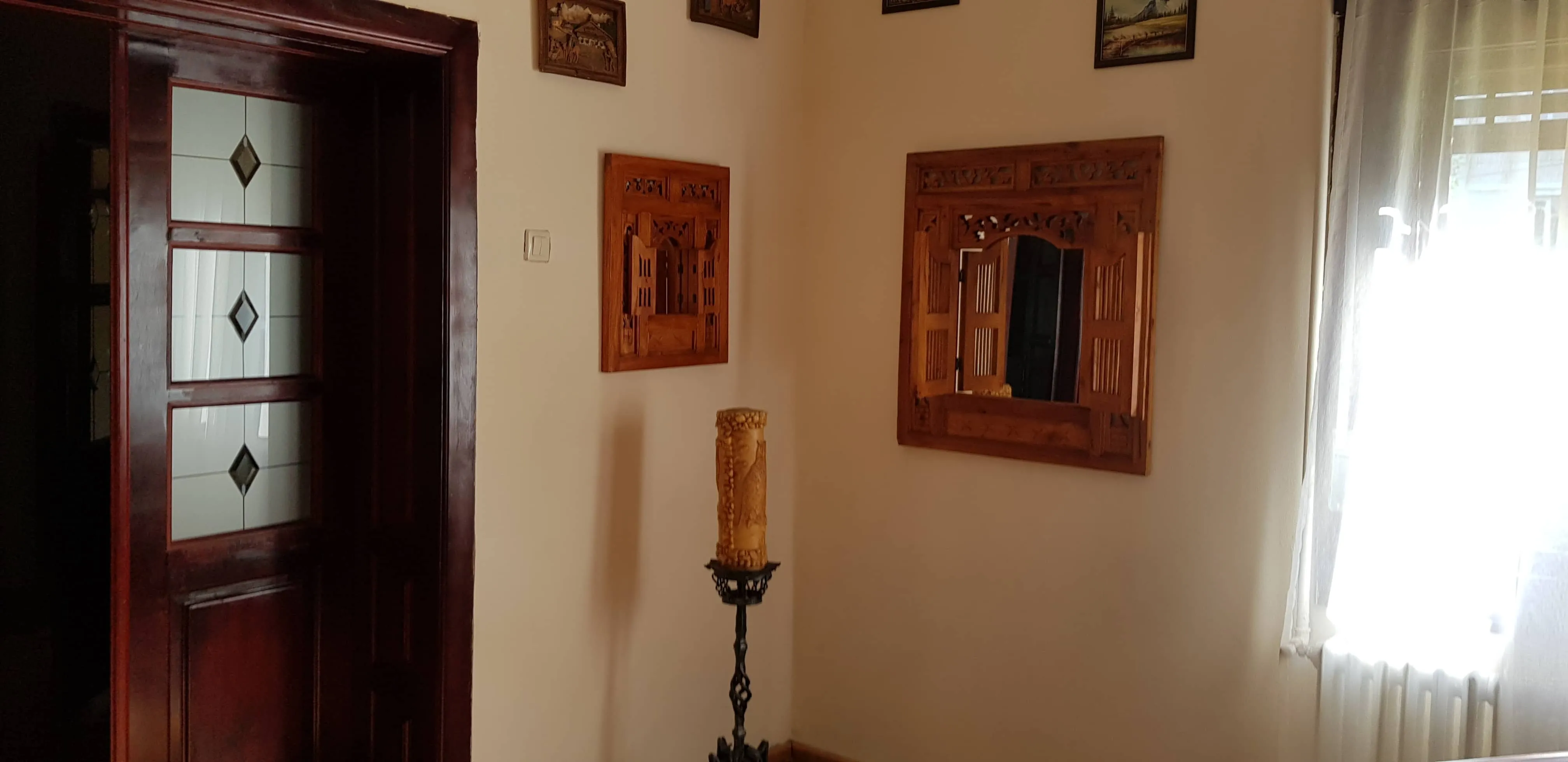 Interior corner of a room with a dark wooden door on the left featuring glass panels with diamond patterns, cream-colored walls, two decorative wooden frames with carved shutters on the wall, a tall ornate candle holder with a carved pillar candle, and a window with sheer white curtains on the right letting in bright light.