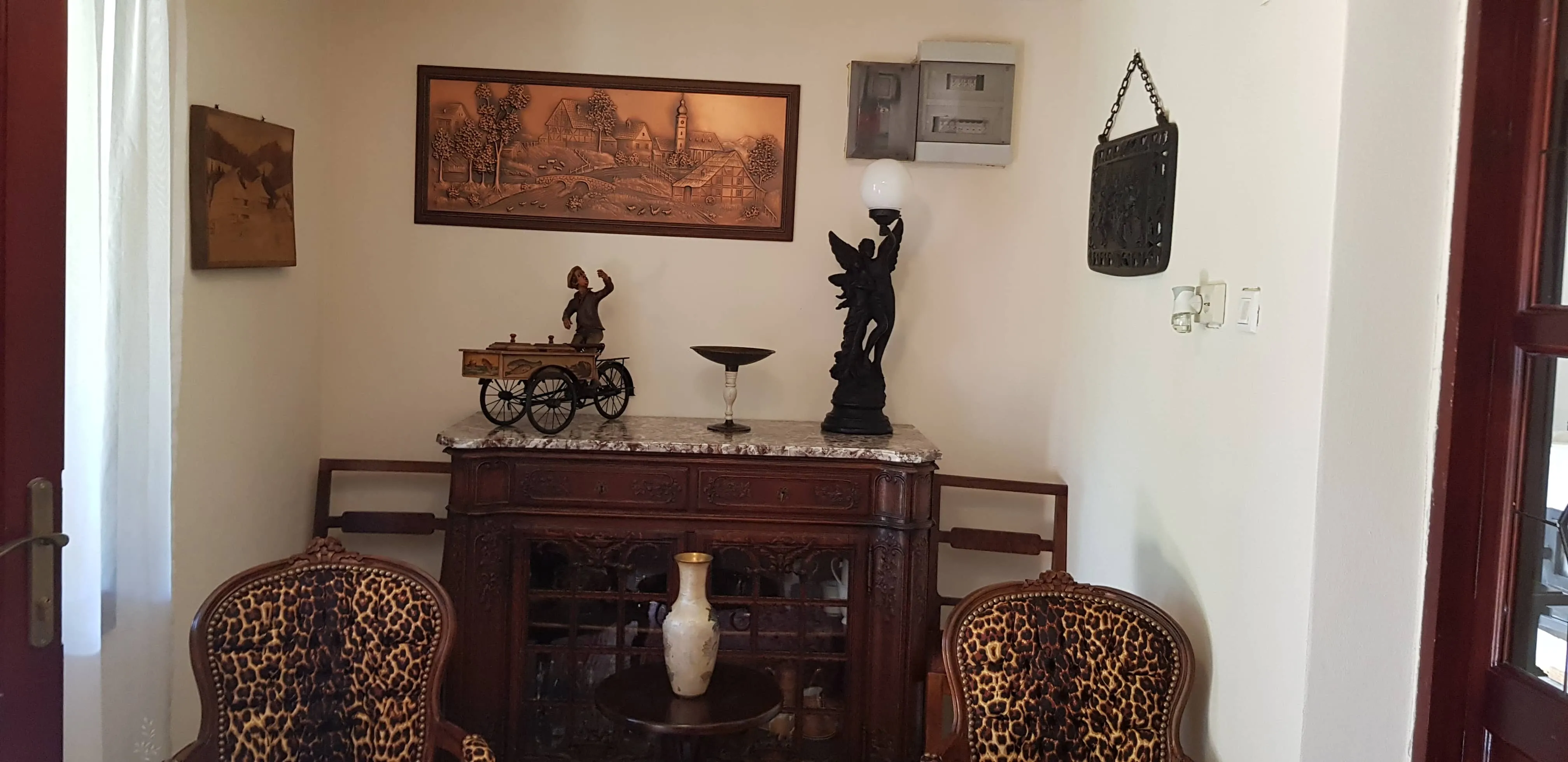 Cozy corner of a room featuring two leopard-print upholstered armchairs, a dark wooden cabinet with a marble top holding a decorative figurine of a man on a tricycle, a pedestal bowl, and an angel lamp, with framed artwork and a wall-mounted decorative piece above.