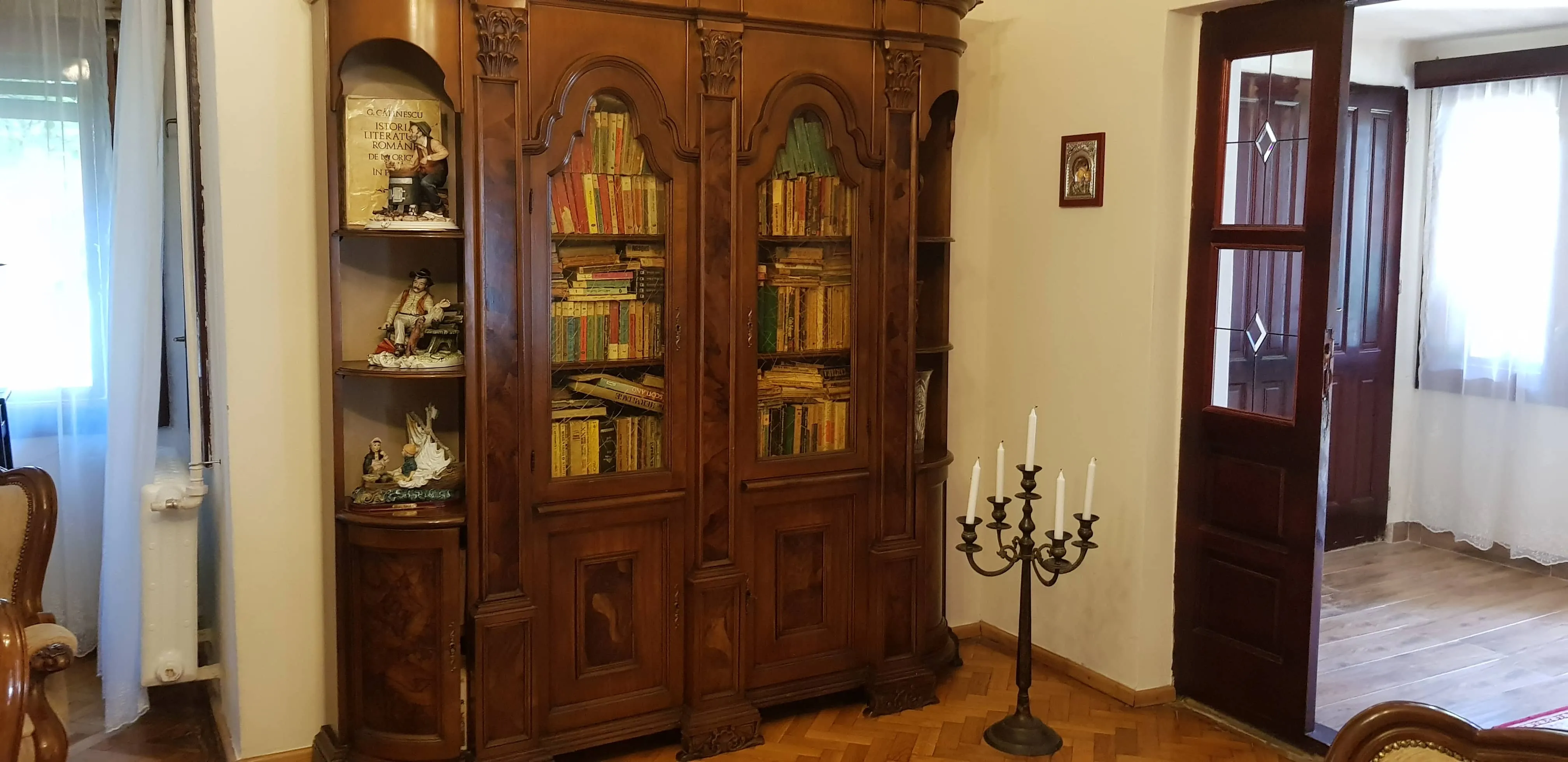 Antique wooden bookshelf with glass doors filled with books, flanked by shelves holding decorative figurines, next to an open door and a metal candle holder with five white candles in a room with parquet flooring.