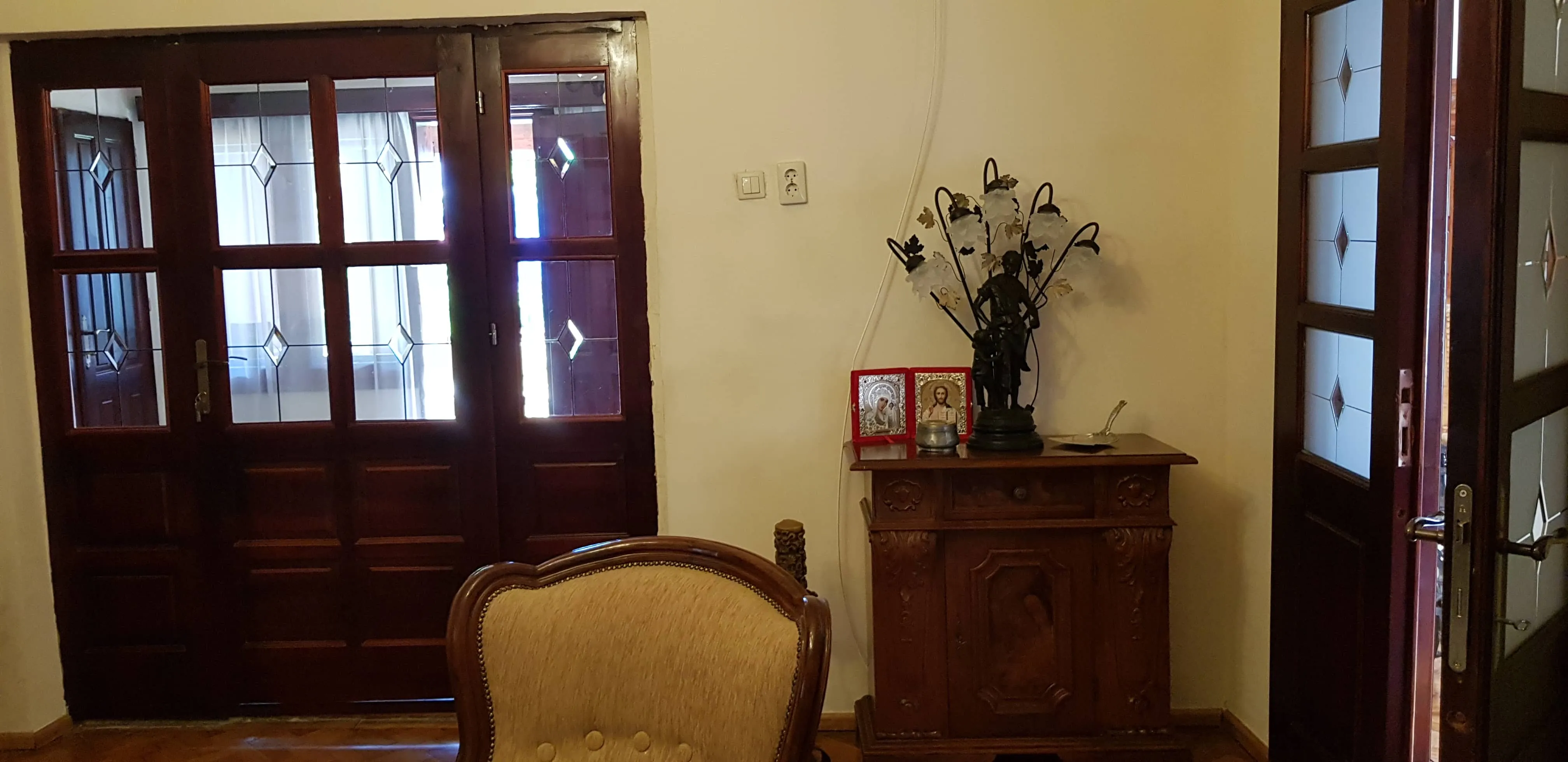 Room interior with dark wooden framed glass double doors on left, an ornate wooden cabinet with decorative lamp and religious icons, and beige cushioned chair in foreground.