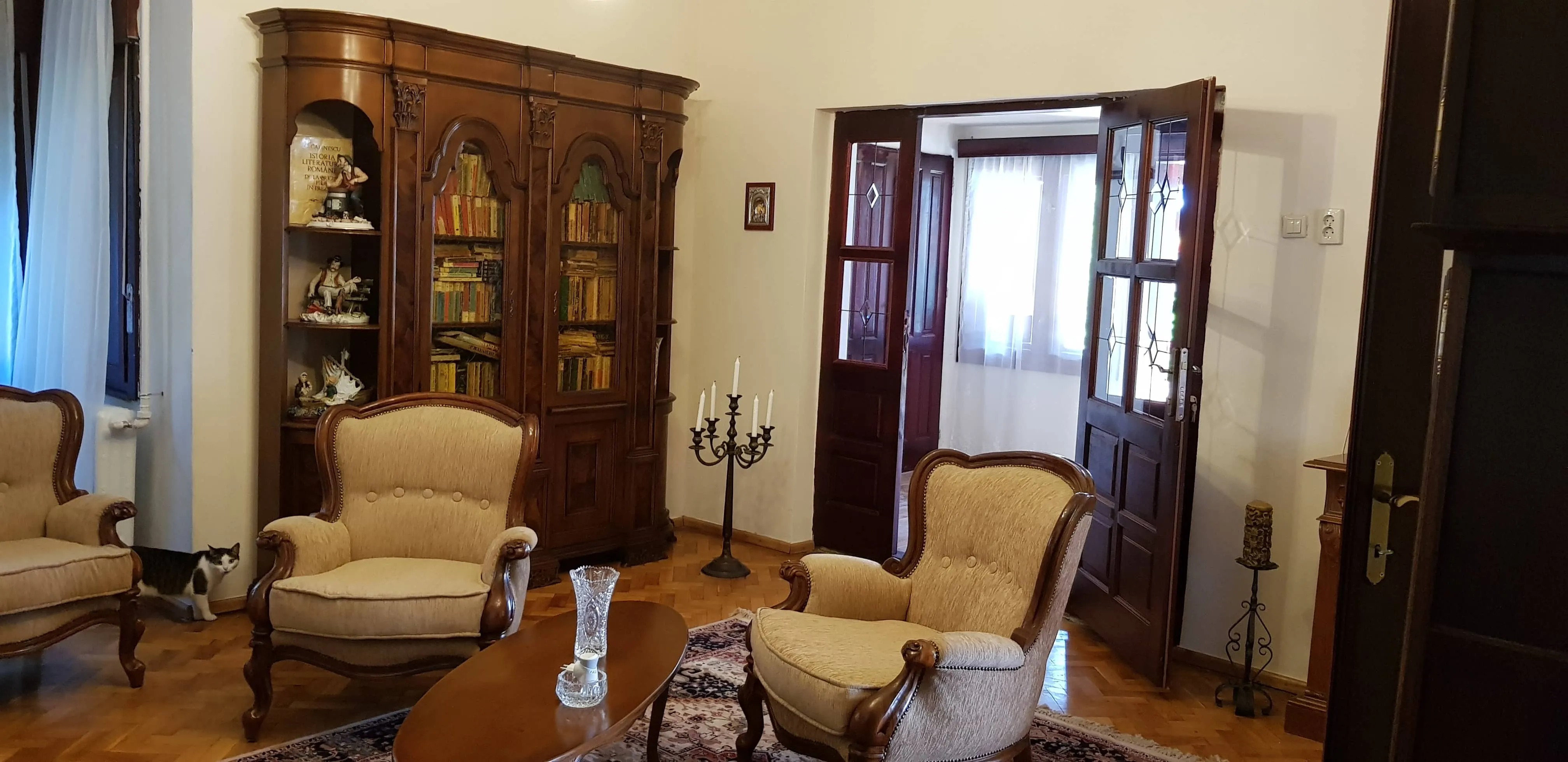 Cozy living room with two beige upholstered armchairs and a wooden oval coffee table holding a crystal vase and candle holder, a large wooden cabinet filled with books and figurines, a tall black candelabra, and an open dark wooden door revealing a room with sheer curtains.