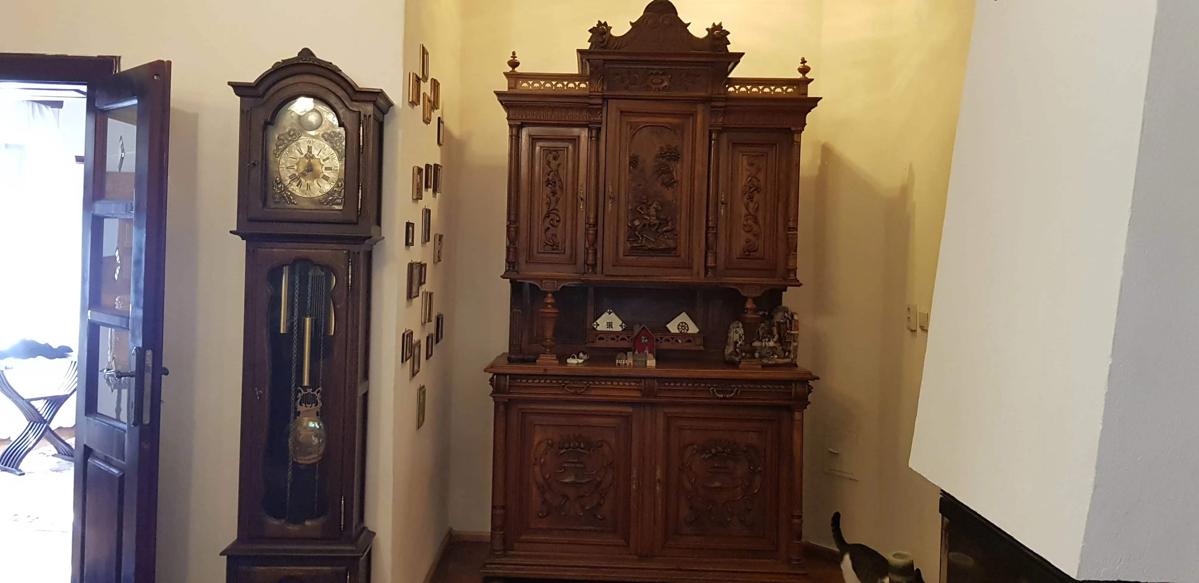Wooden antique grandfather clock with metallic clockface and pendulum stands beside a large ornate wooden cabinet with carved details and miniature house decorations on the shelf, in a room with a partially open door and a cat walking near a fireplace.