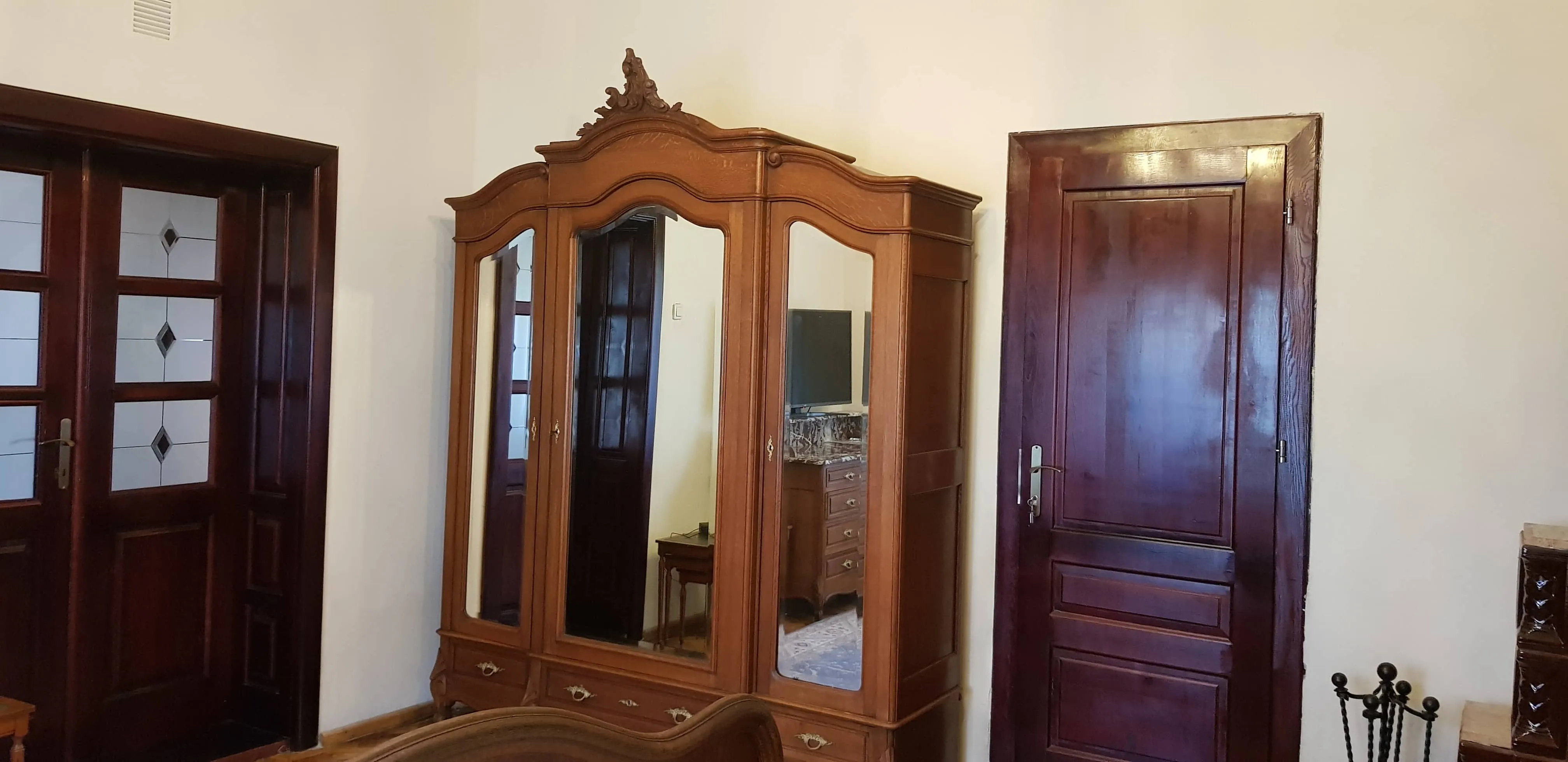 Room interior featuring a vintage wooden wardrobe with three mirrored doors and ornate top carvings, flanked by two dark wooden doors with panels and glass diamond patterns.