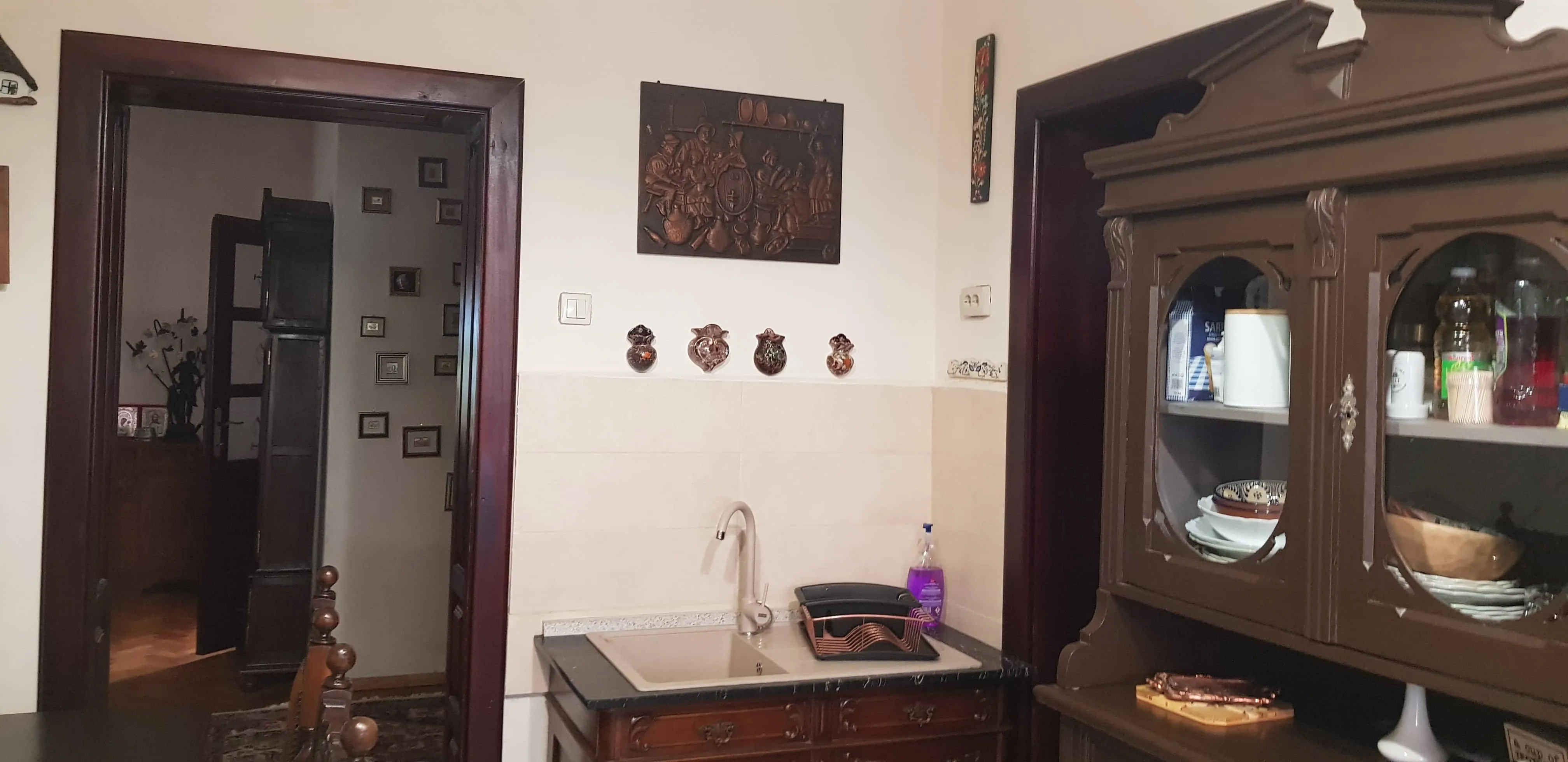 A kitchen corner with a beige sink set in a dark wooden cabinet. Above the sink is a decorative wall hanging and four small ceramic ornaments on the wall. To the right is a dark wooden cabinet with glass doors displaying dishes and kitchen items. A doorway on the left leads to another room with framed pictures on the wall and dark wood furniture.