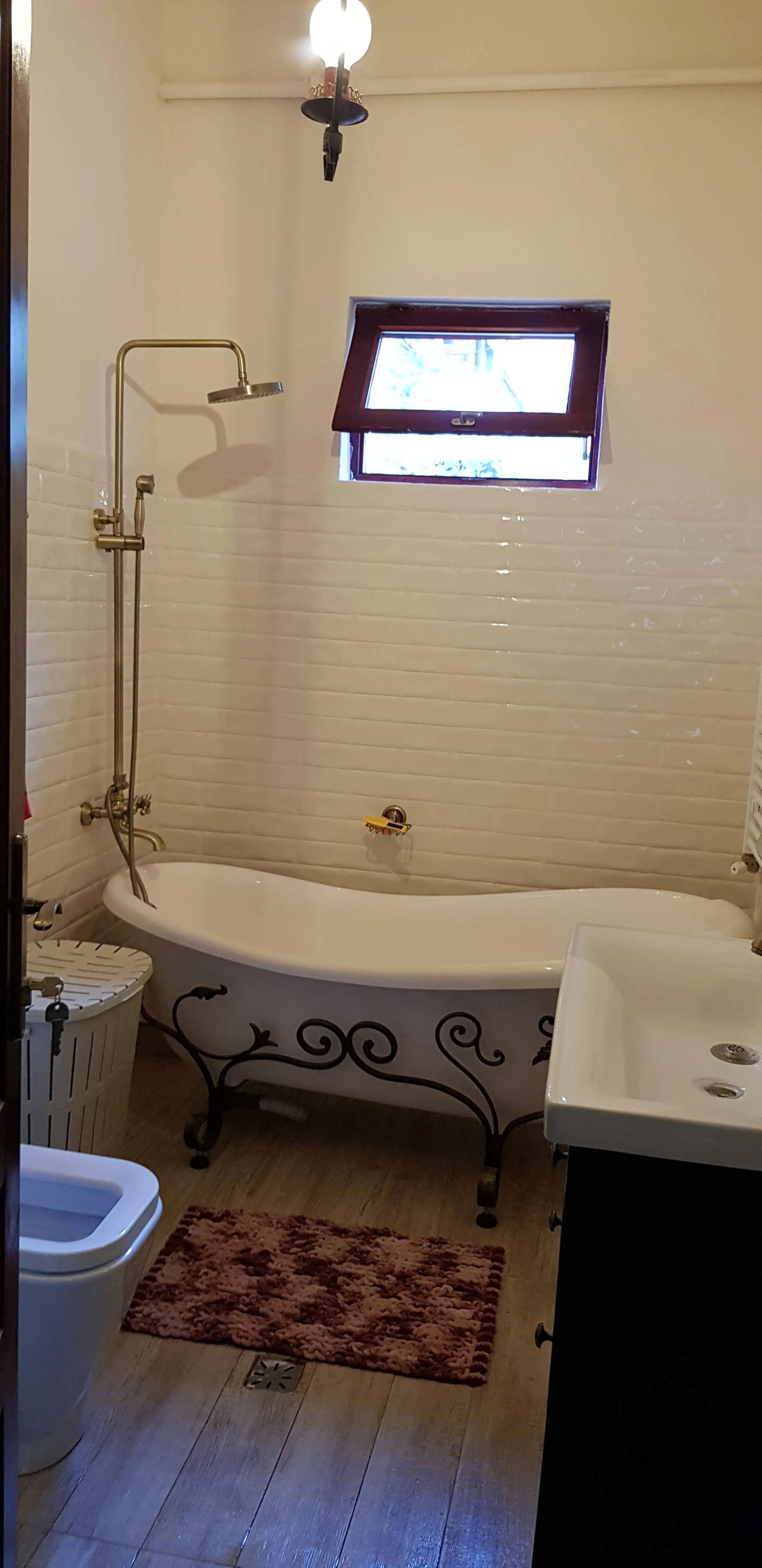 Bathroom with vintage clawfoot bathtub featuring black ornate metal legs, brass shower fixtures on the left wall, a small rectangular window above the tub, white sink on the right, a toilet in the foreground, a tall white laundry basket beside the tub, and a reddish-brown textured bath mat on wood-look floor tiles.