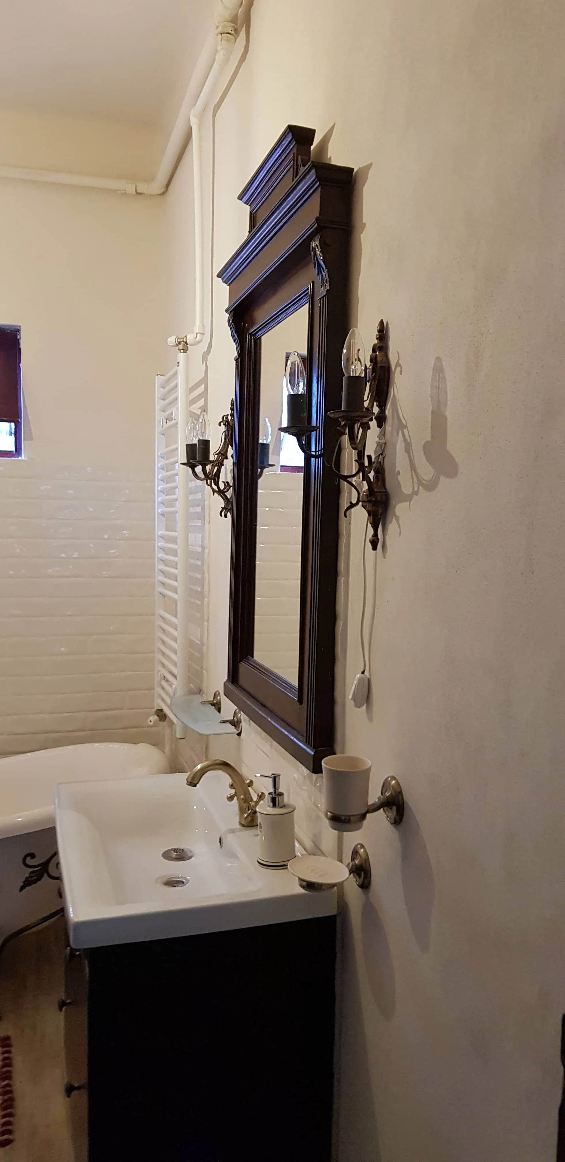 Bathroom interior showing a dark wooden framed mirror with two antique-style wall sconces on either side, a white ceramic sink with brass faucet, soap dispenser, and wall-mounted holder with a cup, white brick-style tiled walls, a white clawfoot bathtub with decorative black feet, and white pipes and radiator on the wall.