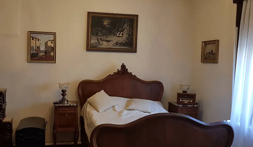 Bedroom with a vintage wooden bed featuring a carved headboard and footboard, white bedding with pillows, two wooden nightstands with decorative lamps, an old-fashioned radio on the right nightstand, and three framed paintings hanging on the walls.
