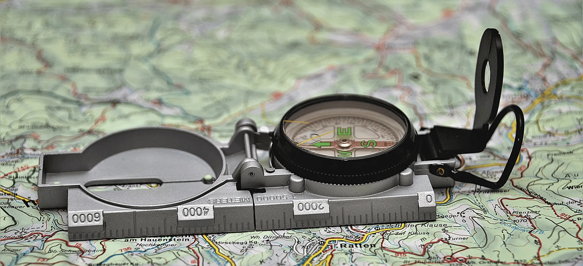 A close-up of a silver and black compass resting on a detailed topographic map with green, red, and yellow markings.