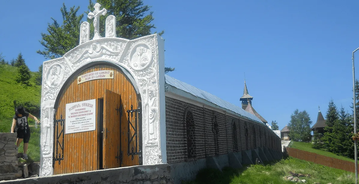 Entrance gate of a wooden and white decorated religious structure with a cross on top, attached to a long brick wall, under a clear blue sky in a green mountainous area.