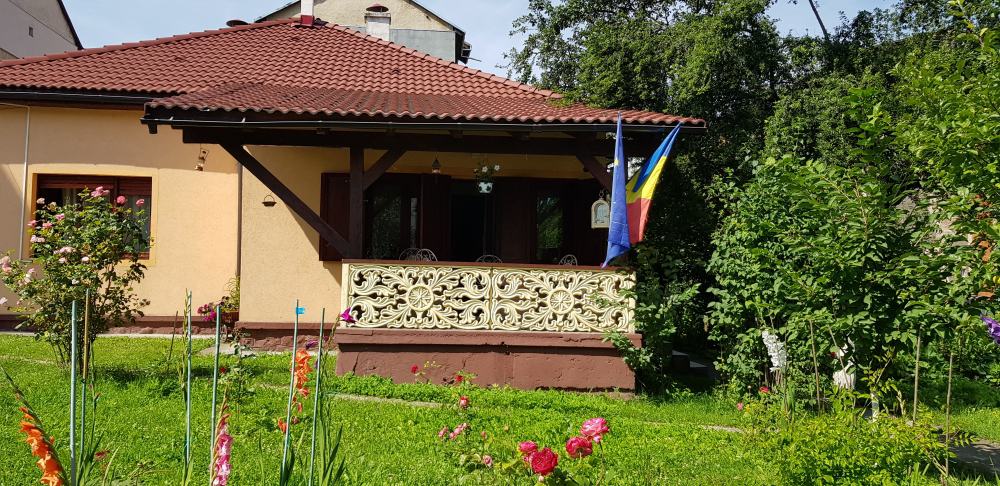 Single-story house with a red tiled roof, a covered porch with decorative balustrade, two flags displayed on the porch, and a garden with green grass, flowers, and bushes in front.