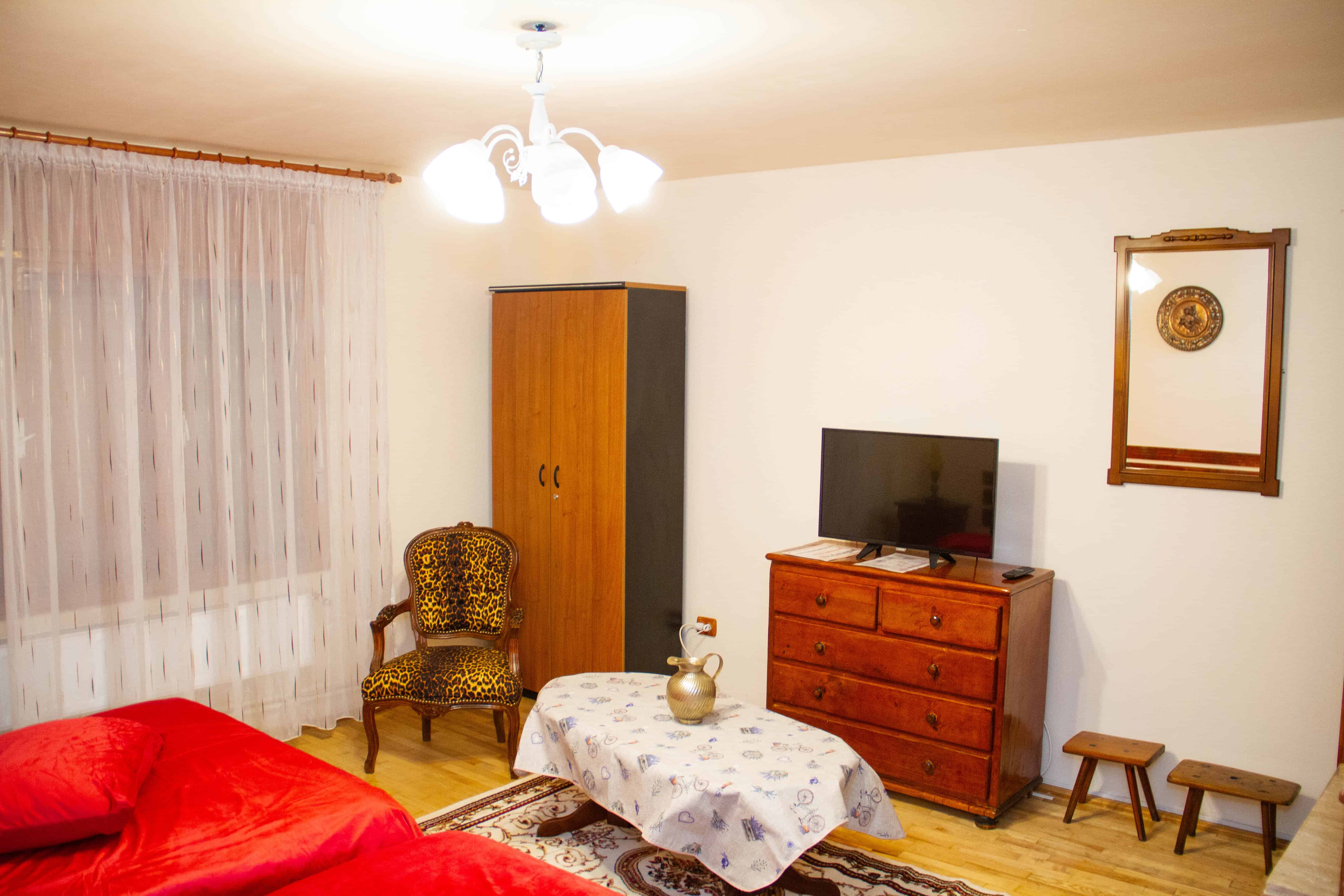 Cozy room with a red bed, vintage armchair with leopard print upholstery, wooden wardrobe, wooden dresser with a flat-screen TV, small wooden stools, table covered with a white cloth and decorative vase, light wood floor, sheer curtain on window, and wall mirror.