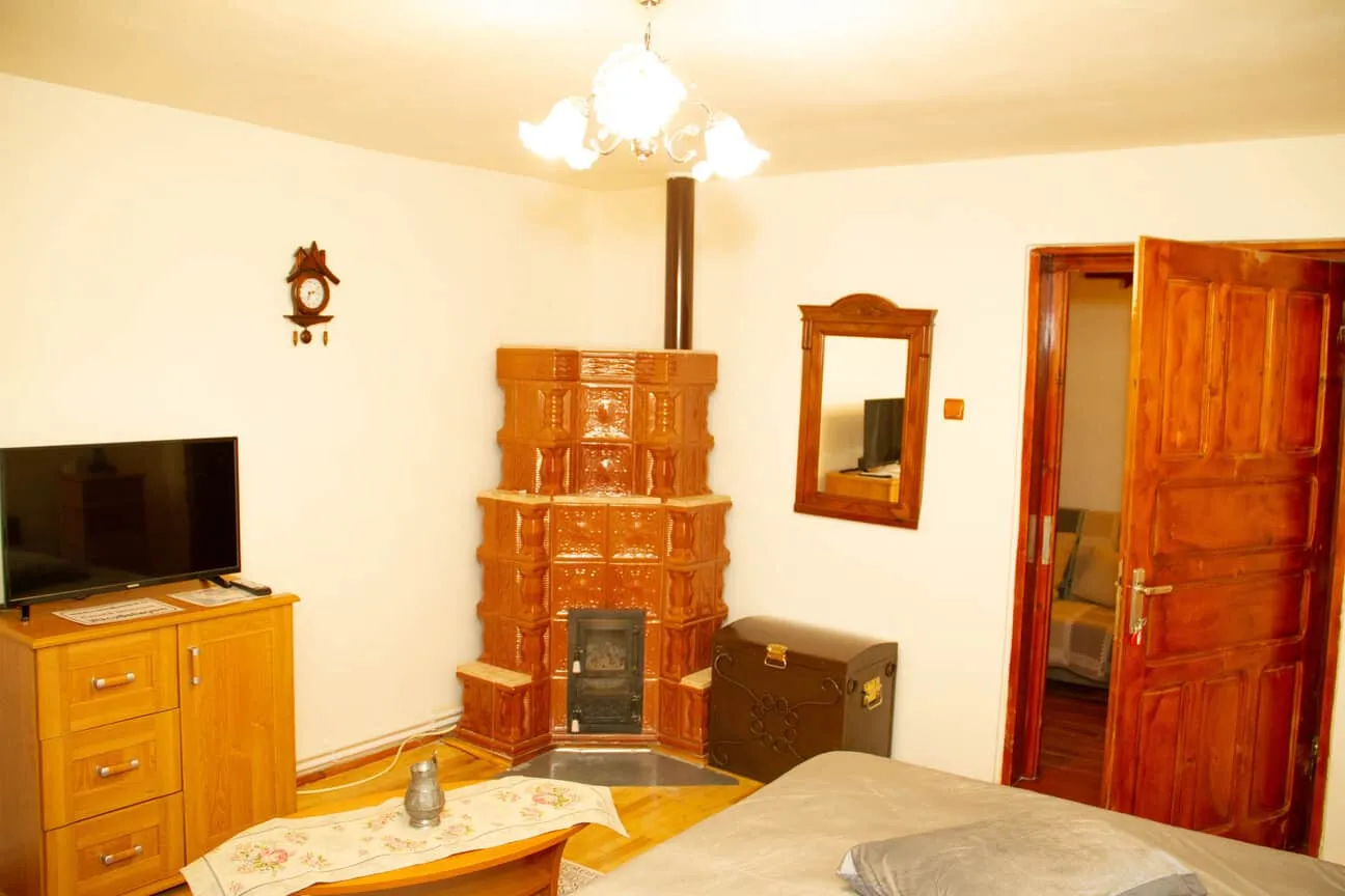 Cozy room with a wooden door, brown tiled corner stove with a chimney, a wooden chest, a wall-mounted mirror, a wooden dresser with a flat-screen TV, a small table with a floral runner and a decorative pitcher, and a bed with a gray blanket.