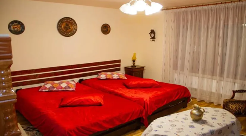A bedroom with two single beds pushed together, both covered with red bedspreads and red pillows, a colorful patterned pillow on each bed, a wooden headboard, a nightstand with a yellow lamp, pale walls with three decorative round wall hangings, a window with sheer curtains, a wooden armchair with patterned upholstery, and a small table covered with a white tablecloth featuring a floral pattern and a brass decorative item on it.