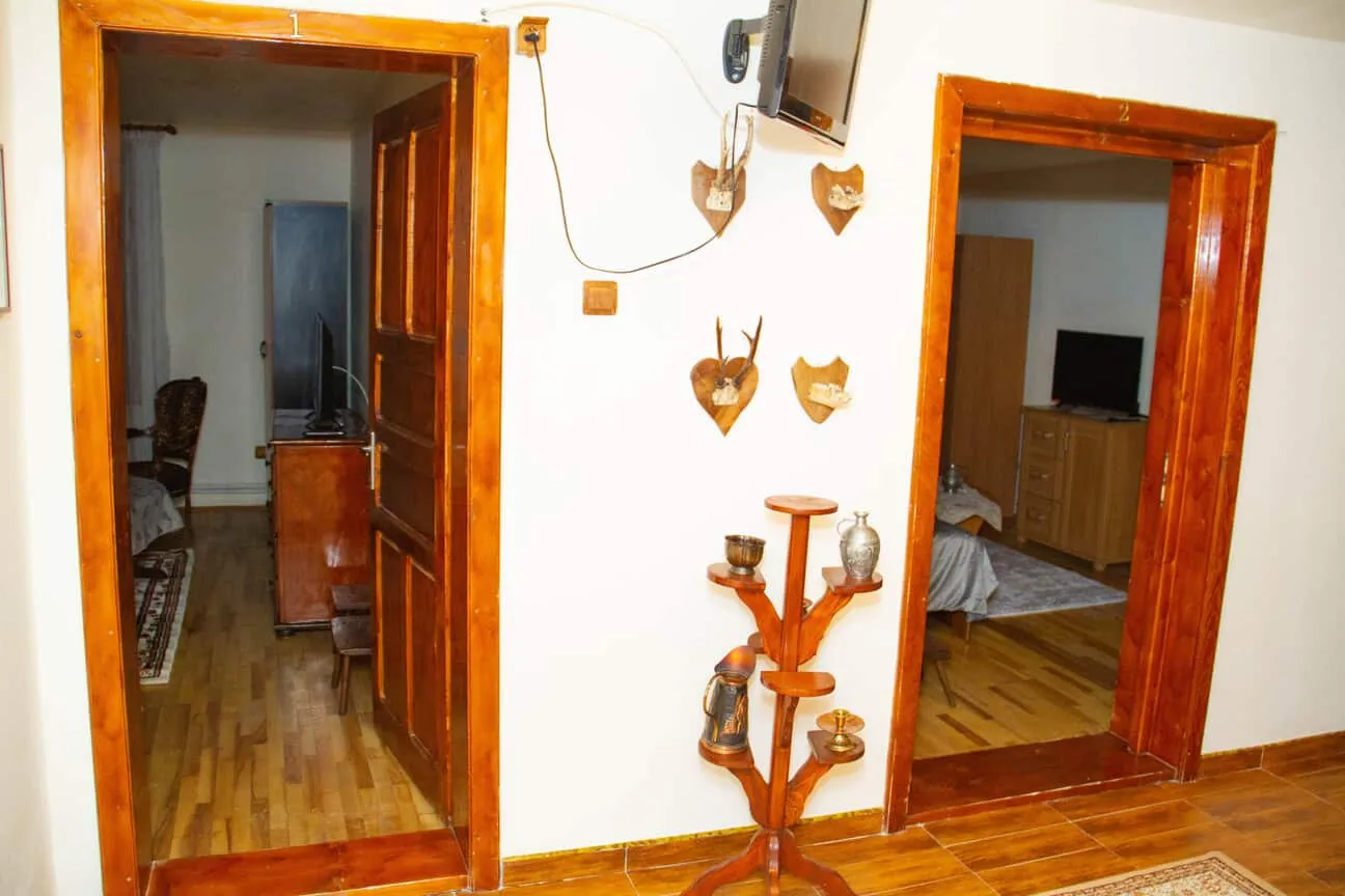 Interior view showing two wooden doorways labeled 1 and 2 leading to rooms with TV sets, a wooden floor, and a decorative wooden stand with various items beneath a mounted TV on the wall.