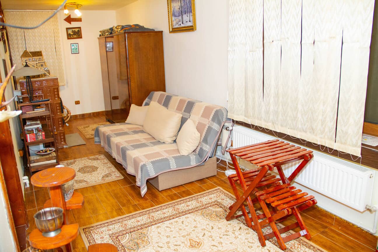 Cozy living room with a checkered sofa covered in a quilt with three beige pillows, a wooden folding table and chair set near the window with white curtains, a dark wooden cabinet, a bookshelf, framed artwork on the walls, and multiple area rugs on a wooden floor.