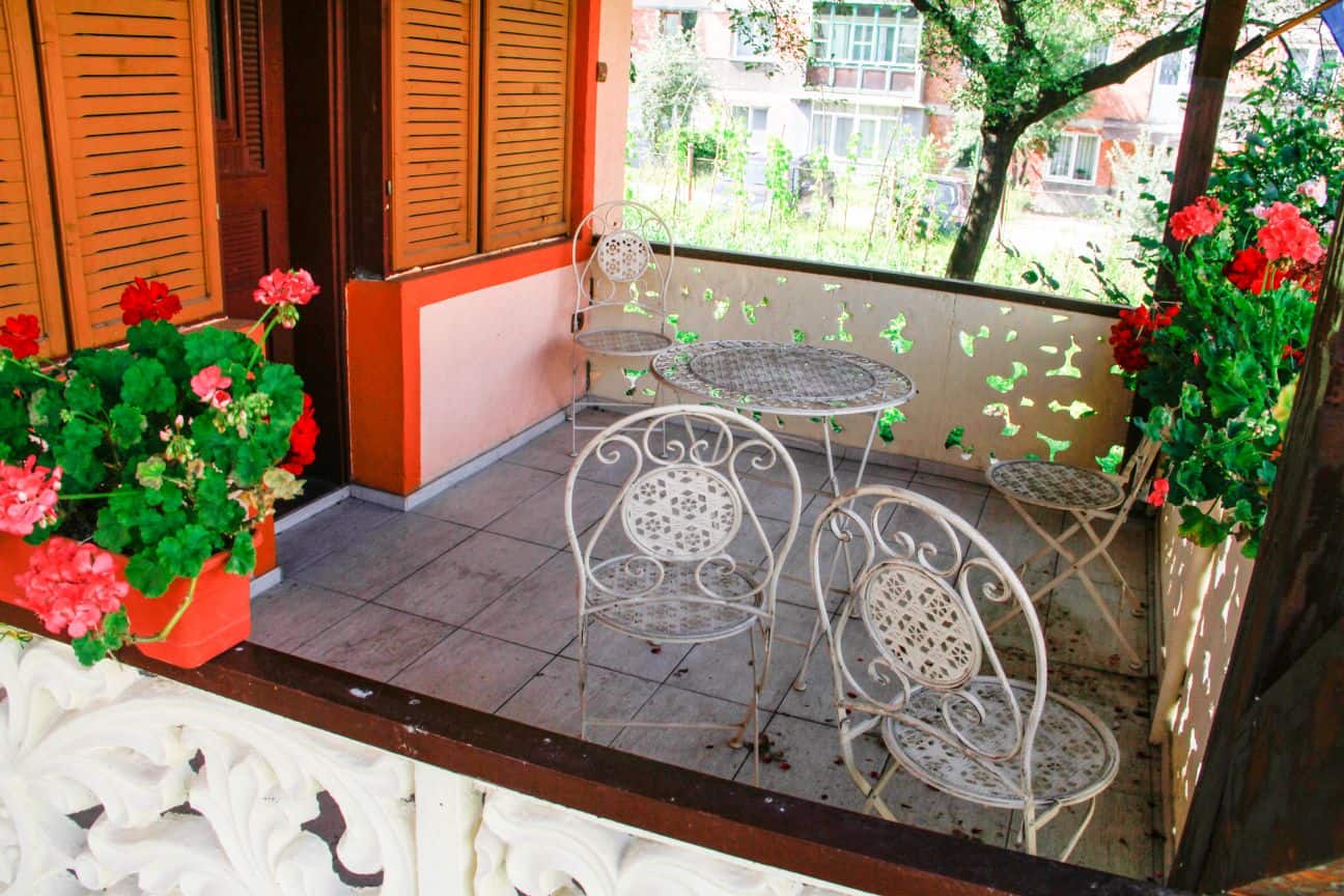 Outdoor tiled patio with white decorative metal table and chairs, surrounded by potted red and pink flowers, with orange shuttered windows and a view of green trees and buildings in the background.
