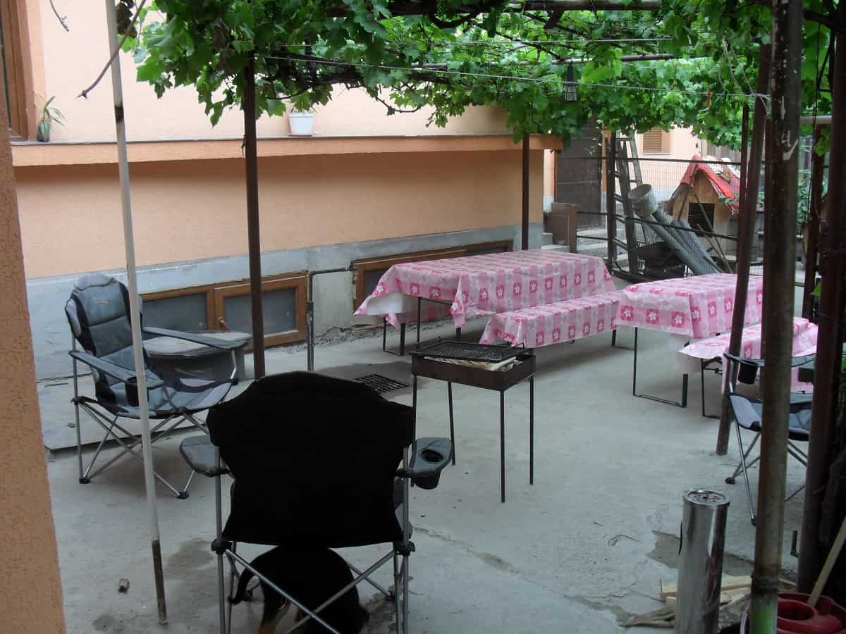 Outdoor seating area under a vine-covered pergola with folding chairs and picnic tables covered with pink floral patterned tablecloths, next to a small metal grill.