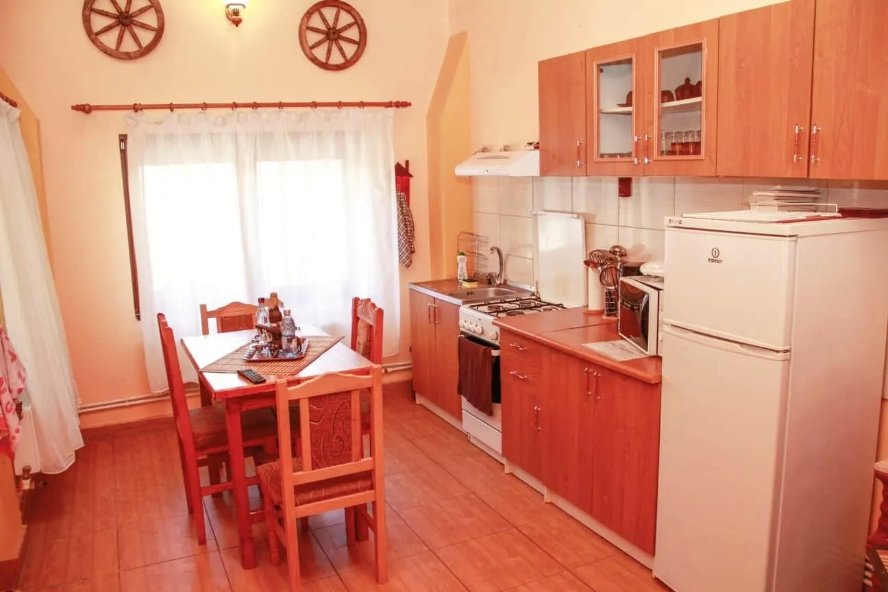 Cozy kitchen with wooden cabinets, a white refrigerator, a gas stove, a microwave on the counter, and a wooden dining table with six chairs near a window with sheer white curtains.