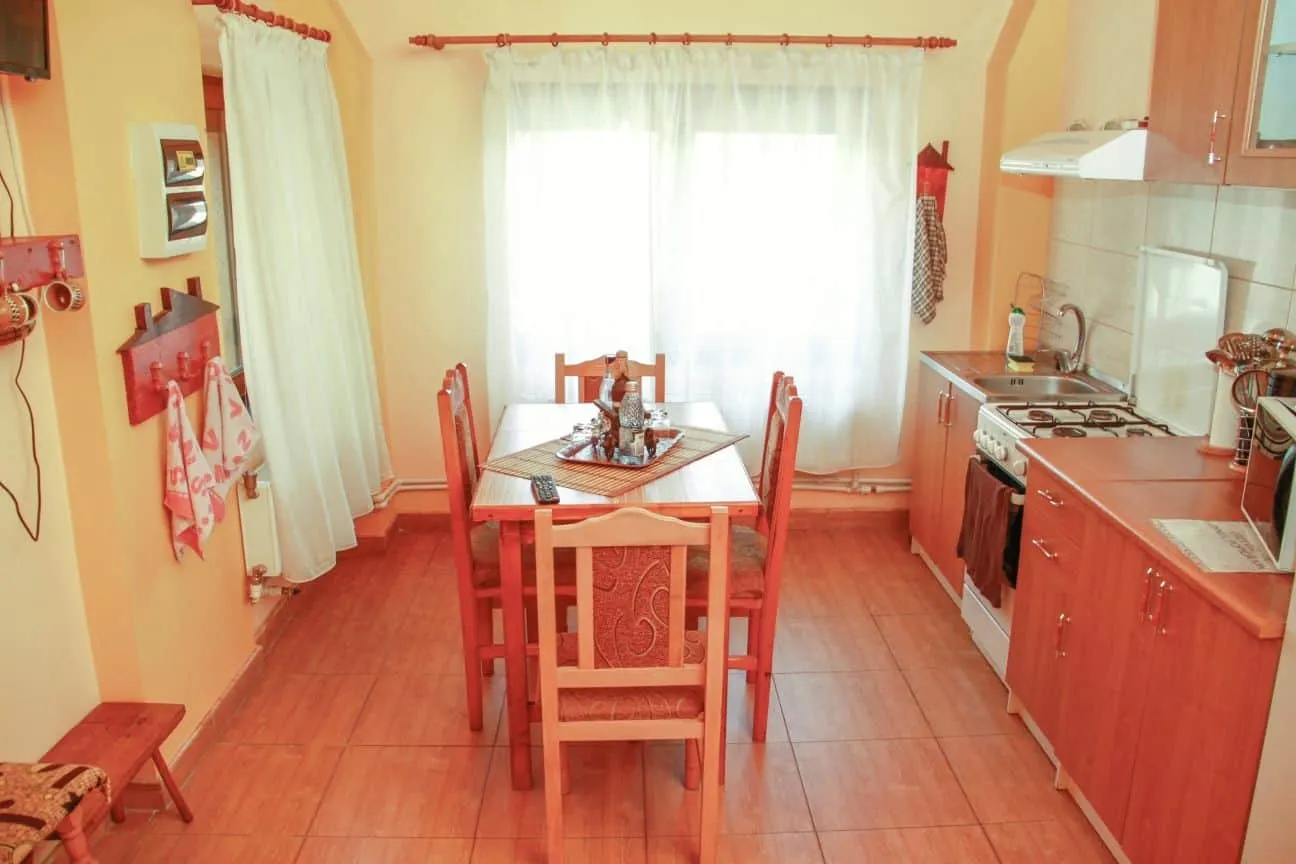 Bright kitchen with wooden cabinets and a gas stove on the right, and a square wooden dining table with six matching chairs in the center beneath a large window covered with sheer white curtains.