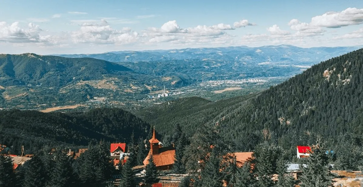 Panoramic view of a mountainous landscape with dense pine forests and scattered houses with red roofs under a cloudy blue sky.