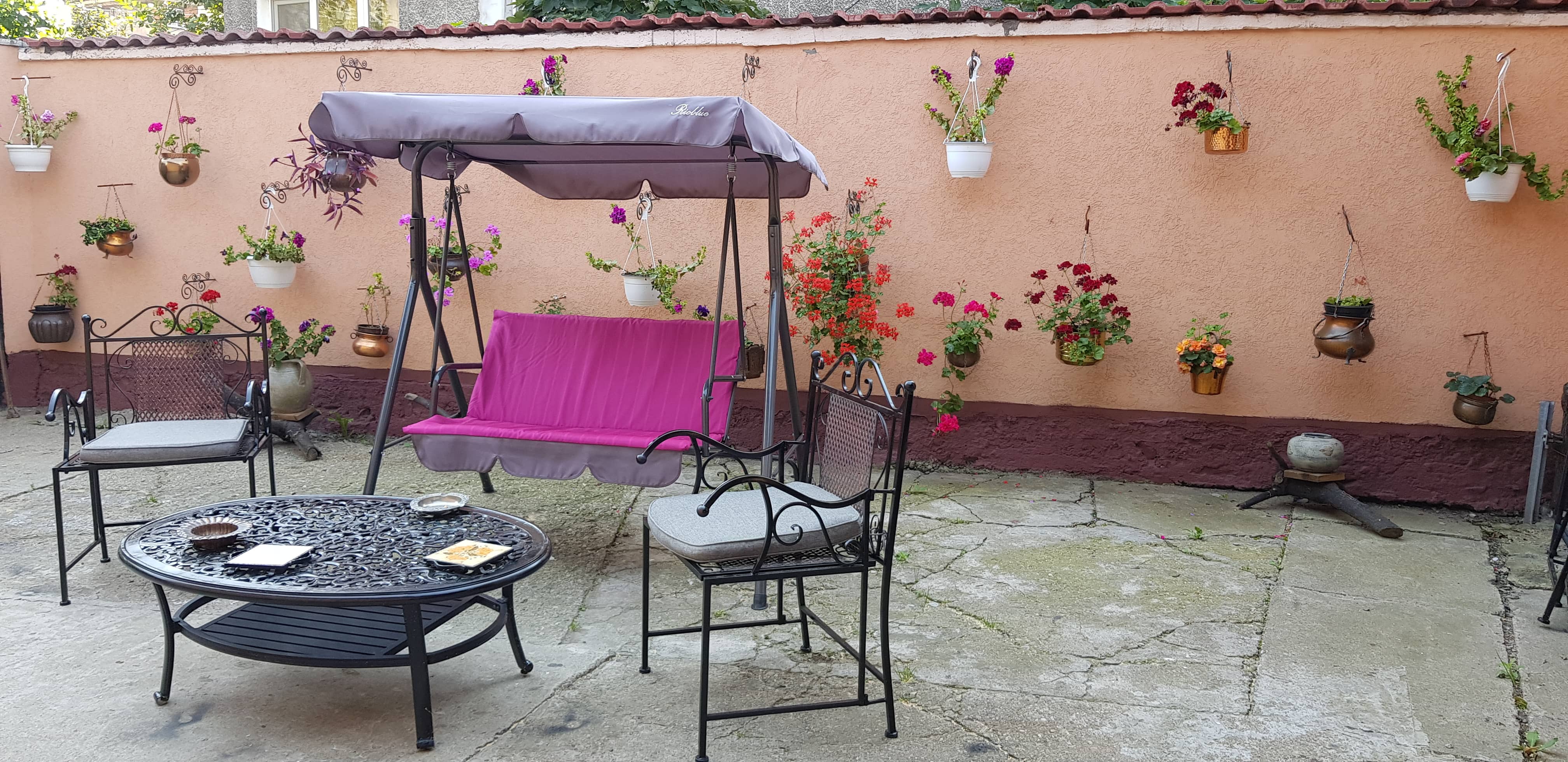 Outdoor patio scene with a black wrought iron round table holding plates and ashtrays, two matching chairs with gray seat cushions, and a purple cushioned swing with a canopy, set against a peach-colored wall adorned with various hanging flower pots with colorful blooms.