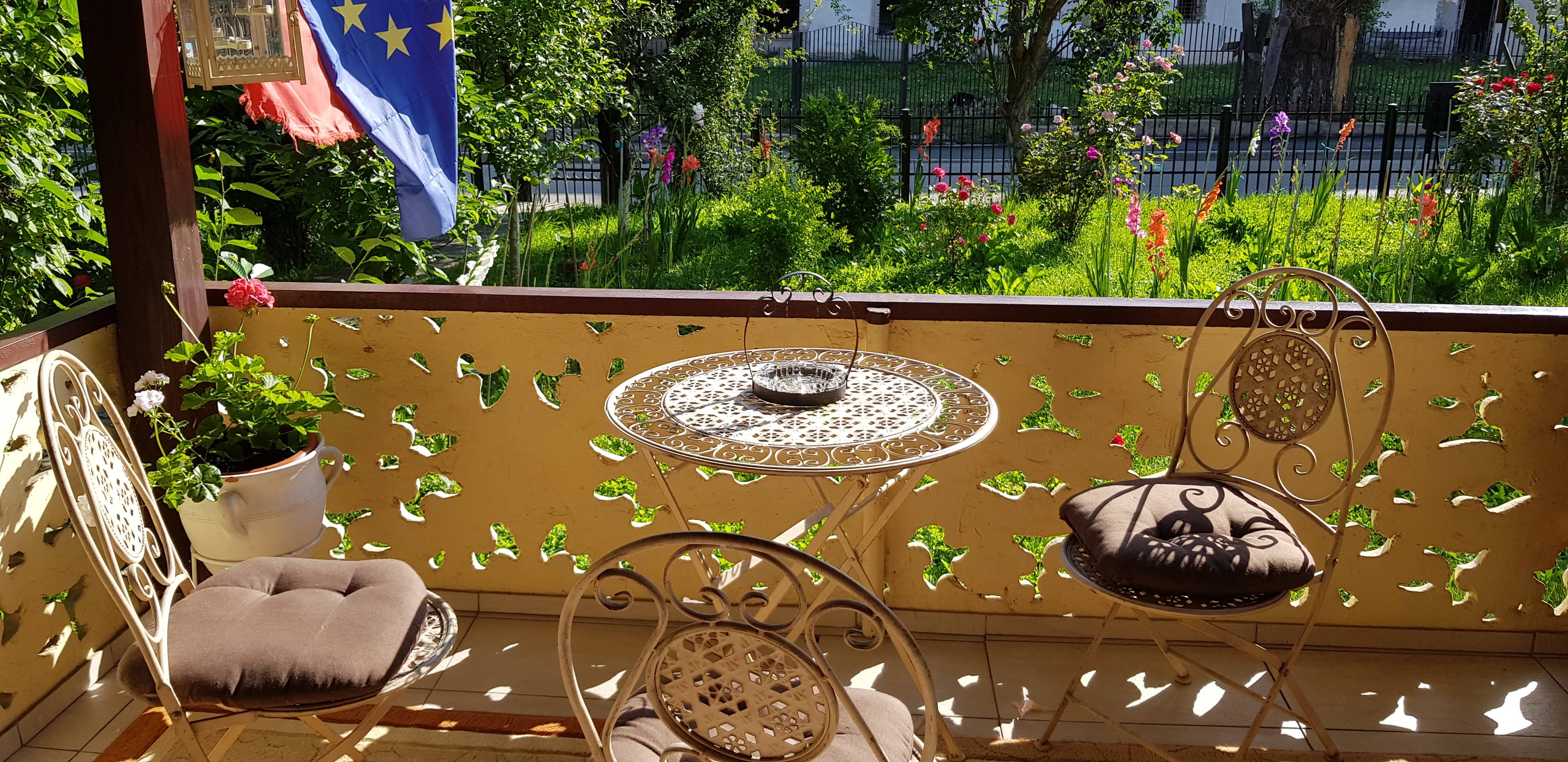 Sunlit outdoor patio with three ornate metal chairs with brown cushions and a matching round table holding a decorative basket, overlooking a green garden with colorful flowers and two flags hanging on the left side.
